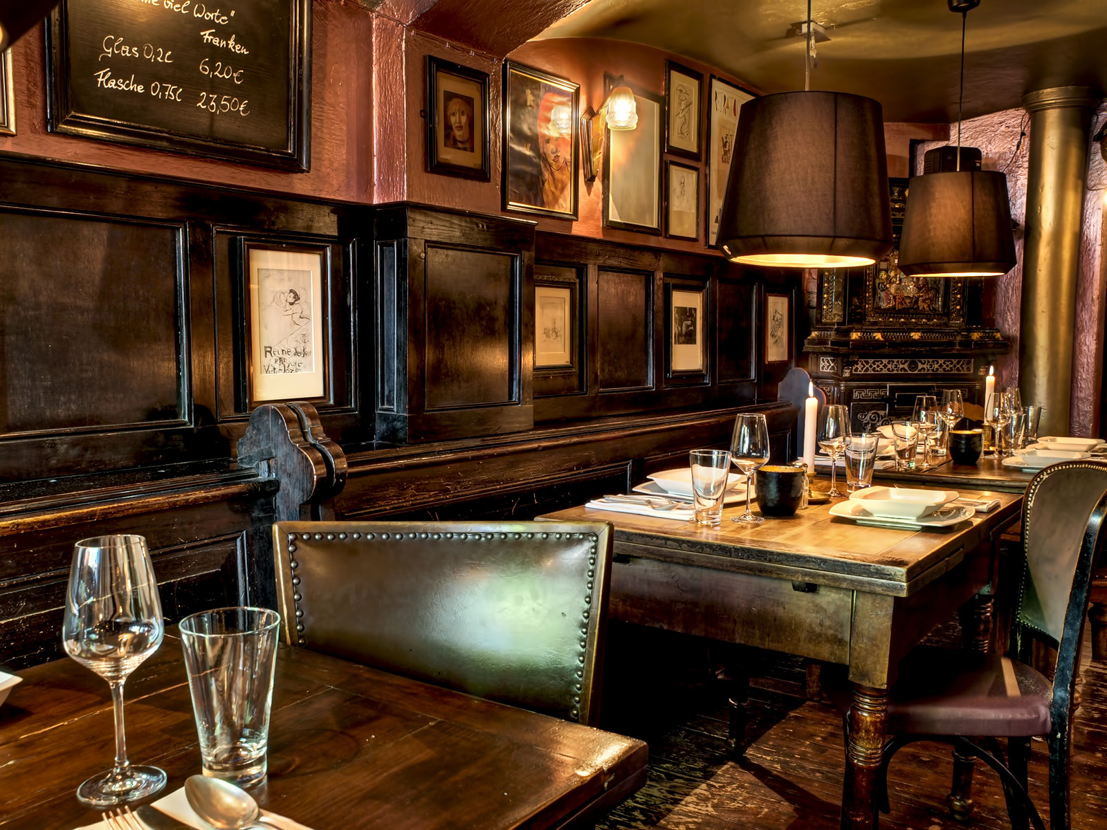 Auf diesem Bild sehen wir einen gedeckten Tisch mit Servietten, Gl&auml;sern, wei&szlig;em Porzellan, Kerzen und silberbesteck. Im Hintergrund befindet sich ein alter Kachelofen im Biedermeier Stil. Dazu sieht man die alten Kirchenb&auml;nke sowie zahlreiche Bilder an den W&auml;nden.