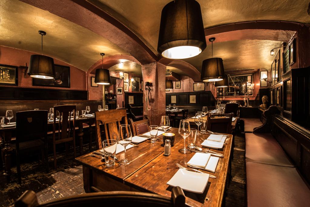 Auf diesem Bild sieht man den vorderen Raum des Restaurants. Im Vordergrund haben wir einen Tisch f&uuml;r vier Personen, dieser ist bereits eingedeckt mit servierten, Besteck, Brottellern, Weingl&auml;sern sowie Wassergl&auml;sern. Man sieht die alte Holzbank, viele Bilder an den W&auml;nden, gro&szlig;e Lampenschirme &uuml;ber den Tischen, die goldene Decke, das Gew&ouml;lbe, die massiven S&auml;ulen.