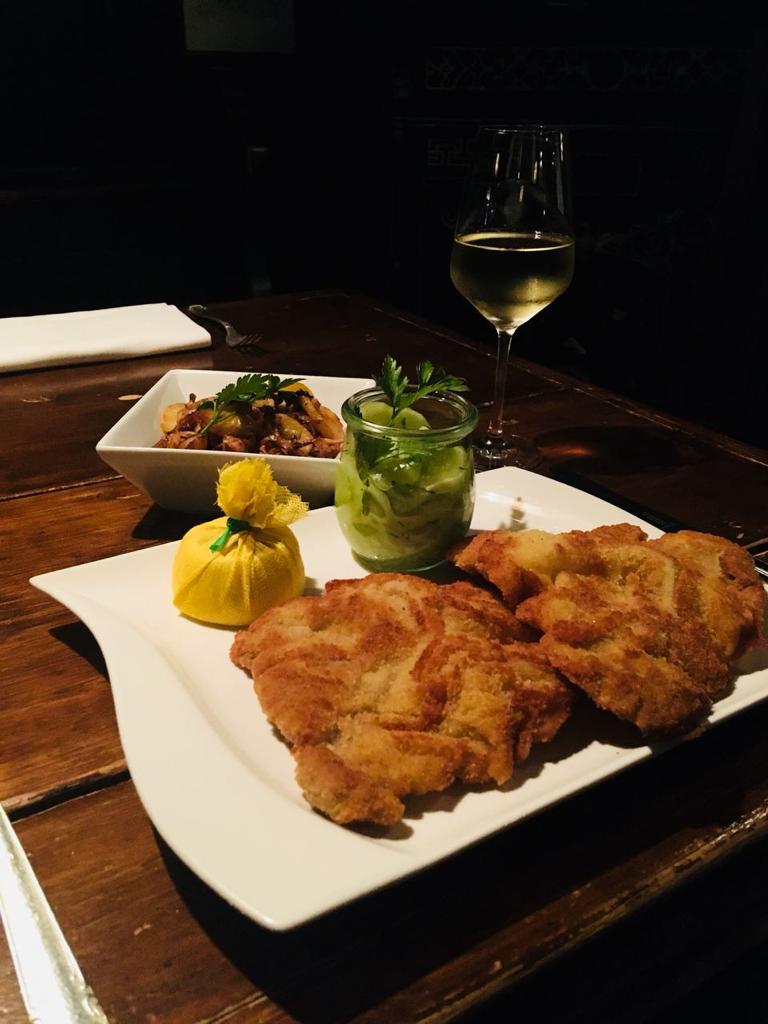 Auf diesem Teller befinden sich zwei appetitlich, braun gebratene Wiener Schnitzel vom Kalb. Dazu ein kleines weg. Glas mit Gurken,salat und eine Zitrone, die in einem kleinen S&auml;ckchen verpackt ist. Dahinter befindet sich ein Sch&auml;lchen mit Bratkartoffeln sowie im anschnitt ein Glas mit Wei&szlig;wein.